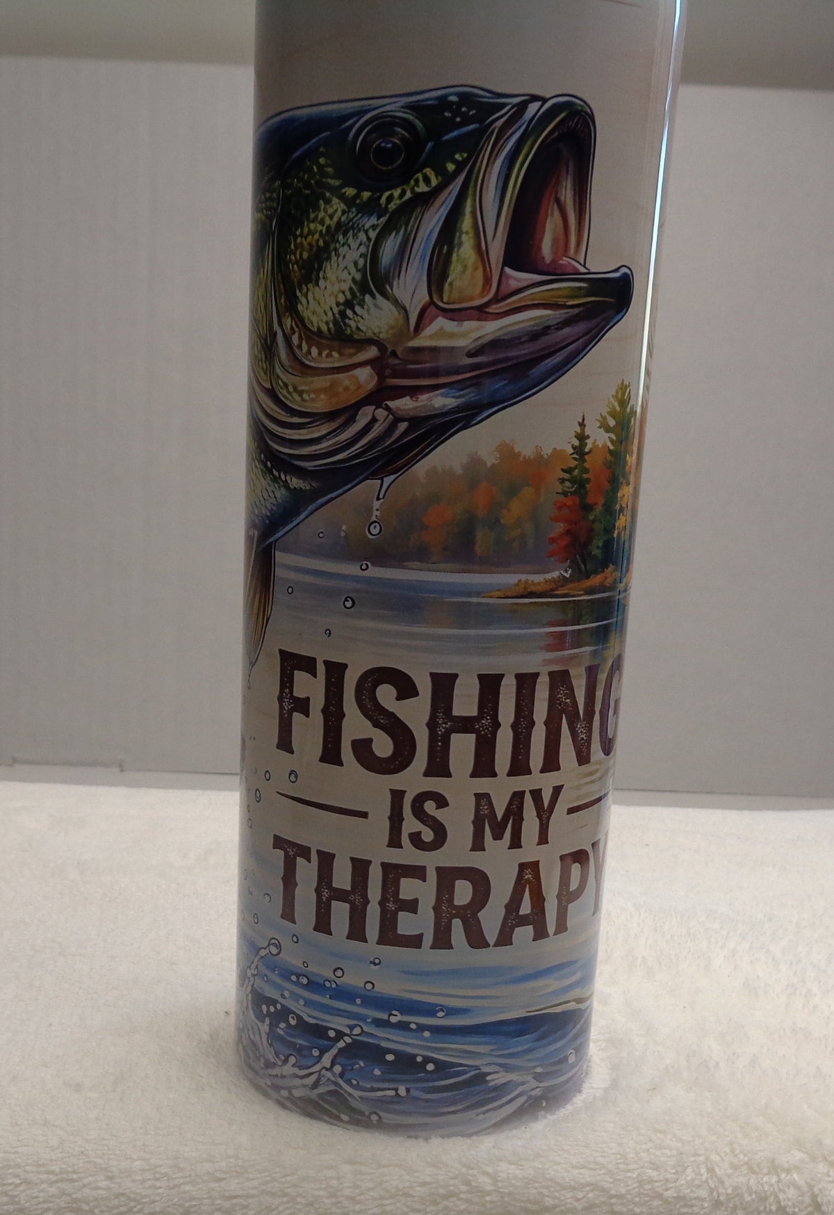 A cylindrical metal can with a fishing-themed label reading 'FISHING IS MY THERAPY' stands upright on a white towel. The label features a large bass fish illustration and blue water graphics.