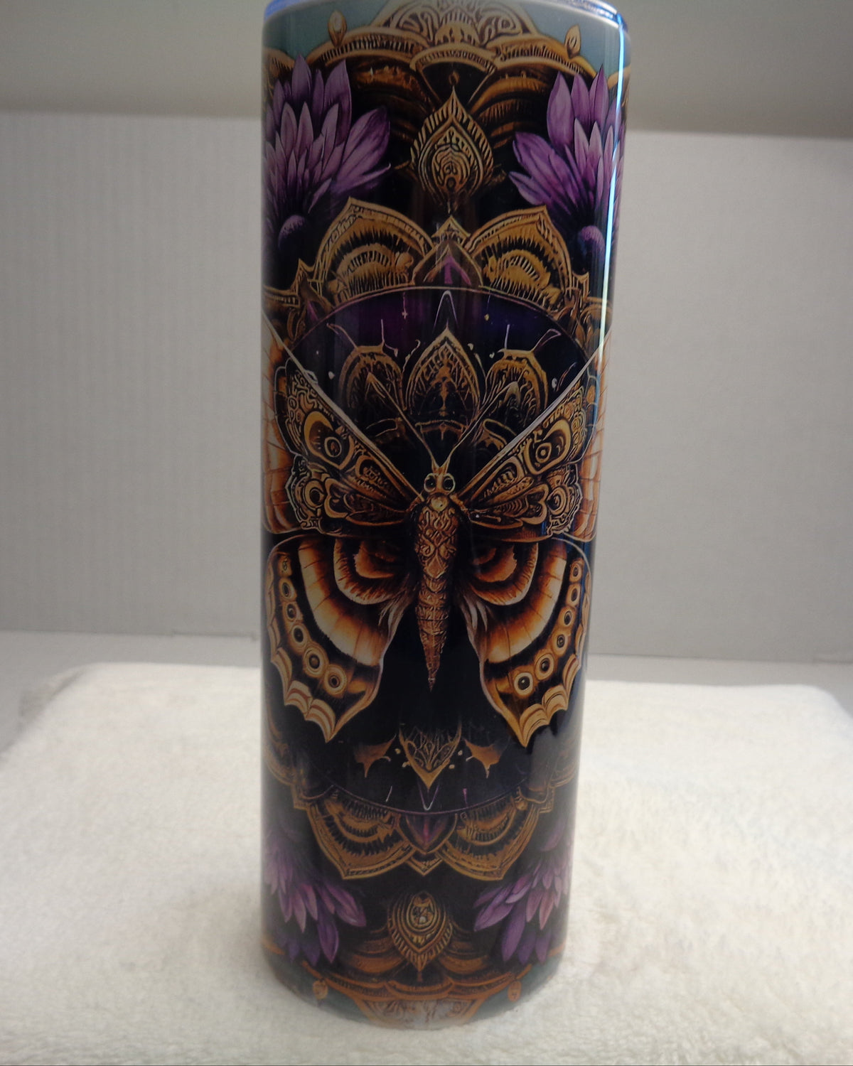 A tall cylindrical tumbler with a detailed symmetrical butterfly and floral design in purple, gold, and black, sitting on a white towel against a white background.