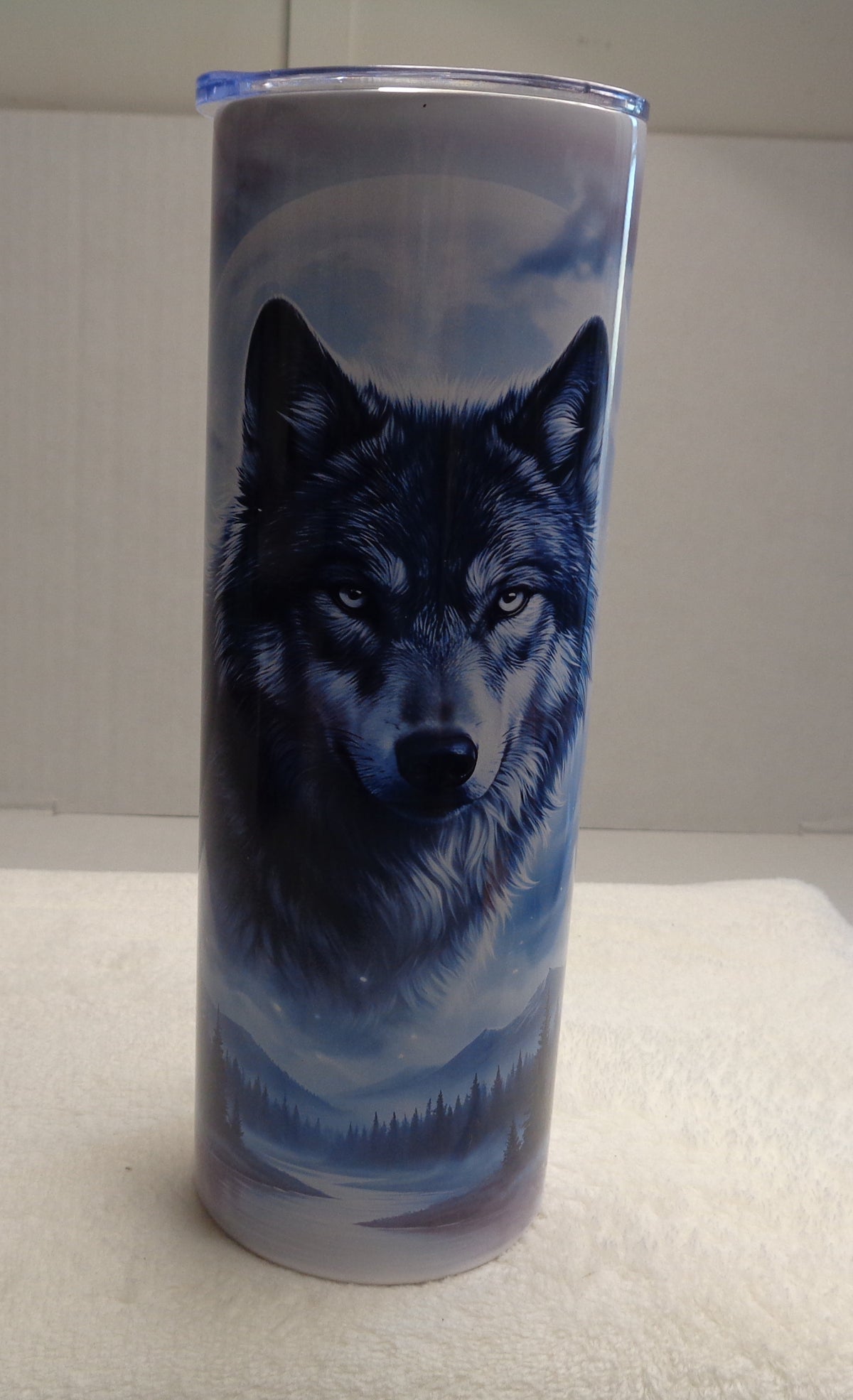 A tall insulated tumbler with a clear lid, featuring a printed graphic of a black and gray wolf against a blue-toned sky and misty landscape. The tumbler is standing upright on a white textured surface against a plain light gray background.