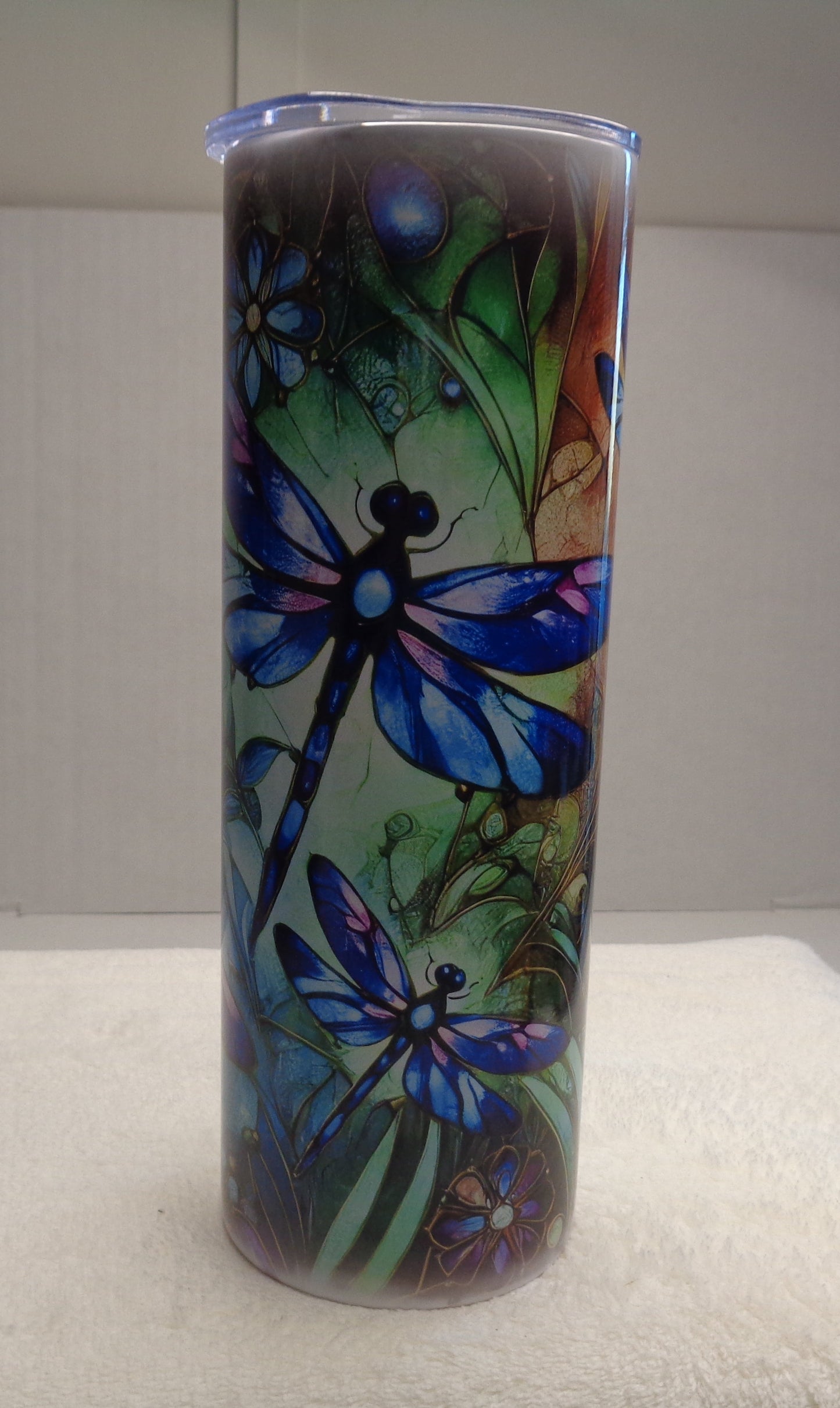 A tall cylindrical tumbler with a clear lid, decorated with a colorful dragonfly and floral pattern, standing on a white towel against a white background.