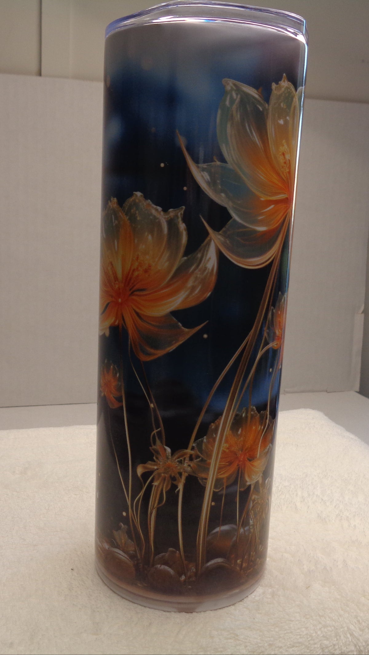 A tall cylindrical glass jar candle with a clear lid, decorated with a dark navy blue background and large orange and yellow floral designs, standing on a white textured surface.