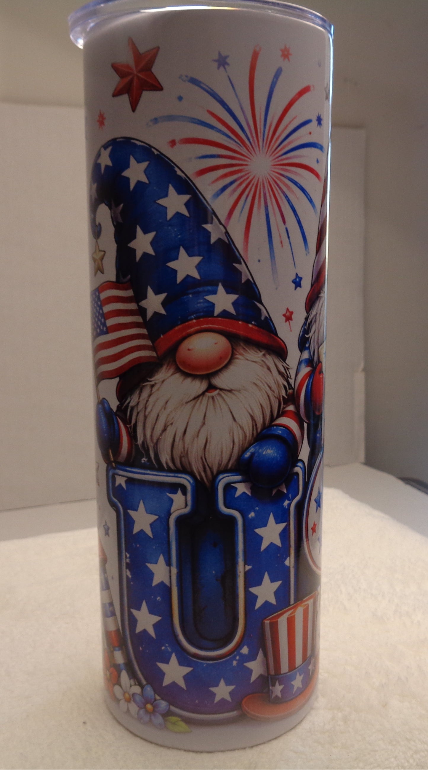 A tall white tumbler with a patriotic 4th of July gnome design, featuring a gnome in a blue star-spangled hat and a large letter 'U' decorated with stars and stripes, set against a home background.