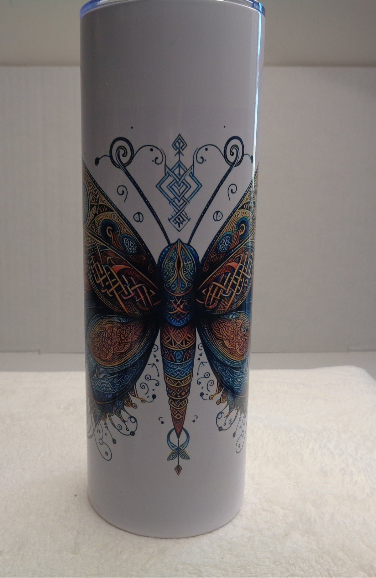 A tall white insulated tumbler with a blue lid, featuring a detailed, colorful mandala-style butterfly design on the front, set against a plain indoor background.