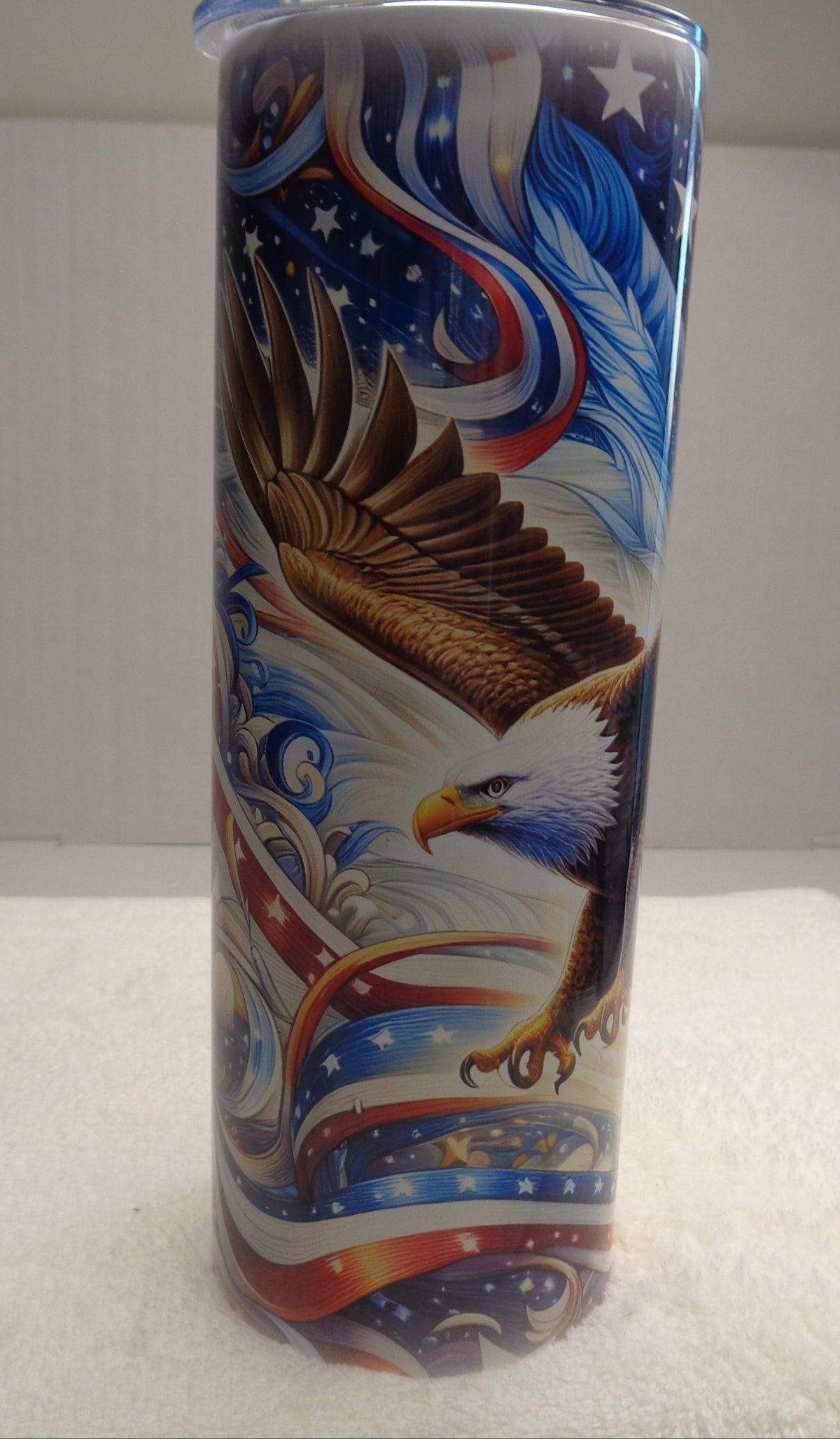 A tall cylindrical tumbler with a patriotic American flag design featuring a bald eagle, blue and white waves, and stars, displayed on a white towel against a white background.