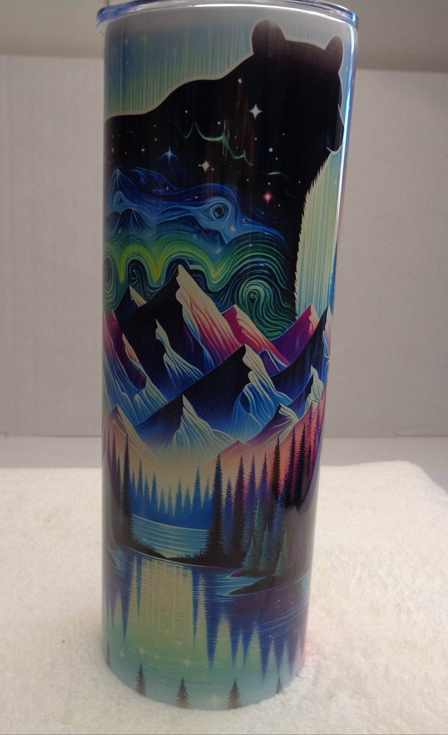 A tall insulated tumbler with a colorful printed design featuring a black bear silhouette, mountains, aurora-like waves, and a lake reflection, shown standing upright on a white surface against a light gray background.
