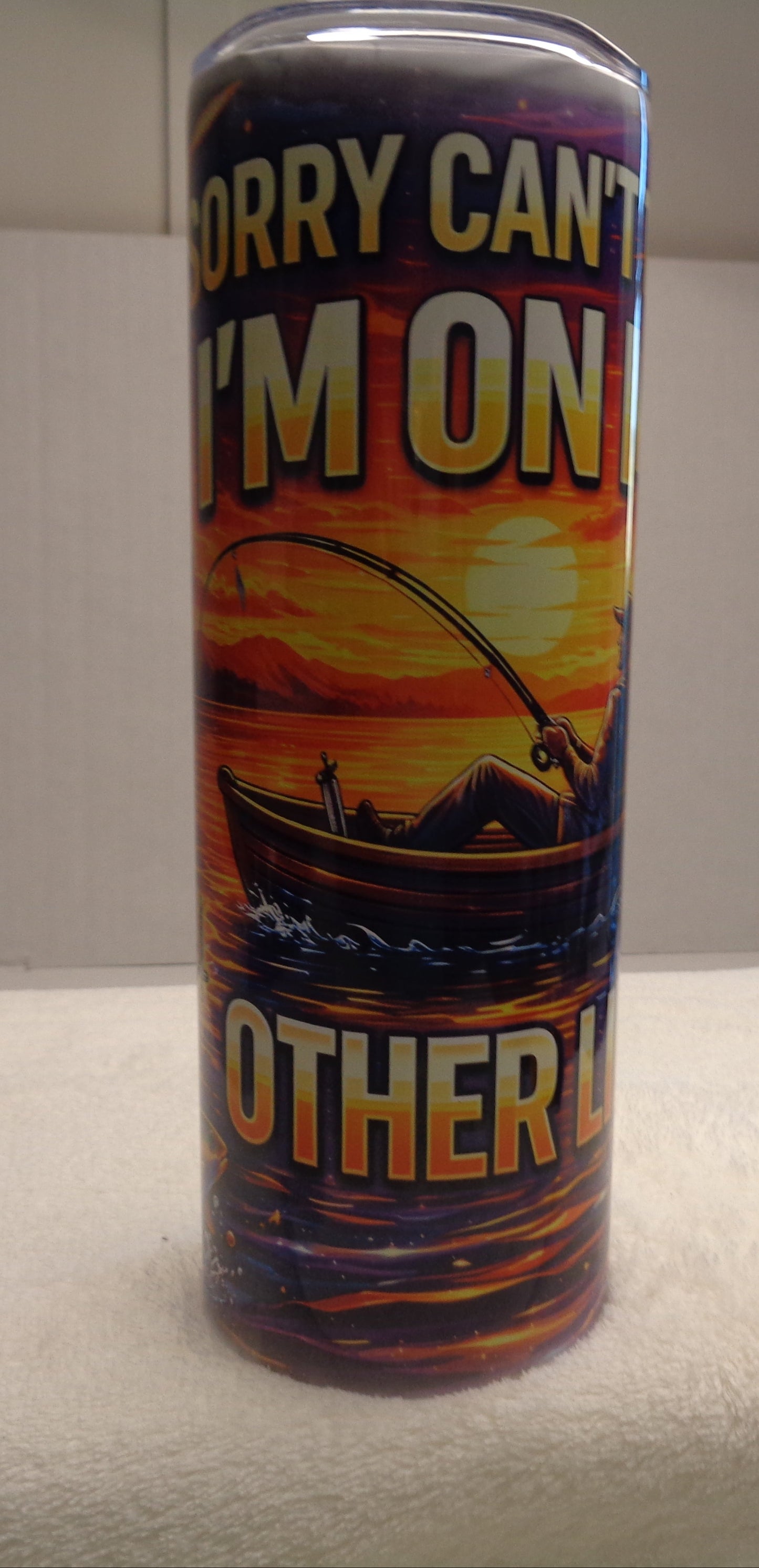 A cylindrical tumbler with a fishing-themed wraparound print, featuring a sunset scene with a boat and the text 'SORRY CAN'T I'M ON OTHER L', displayed on a white towel on a tabletop.