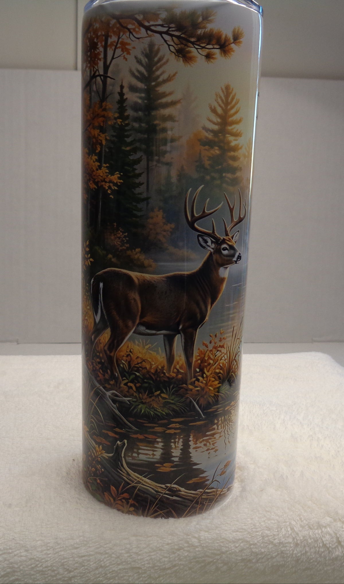 A tall cylindrical insulated tumbler with a detailed deer in a forest design, standing on a white towel against a white background.