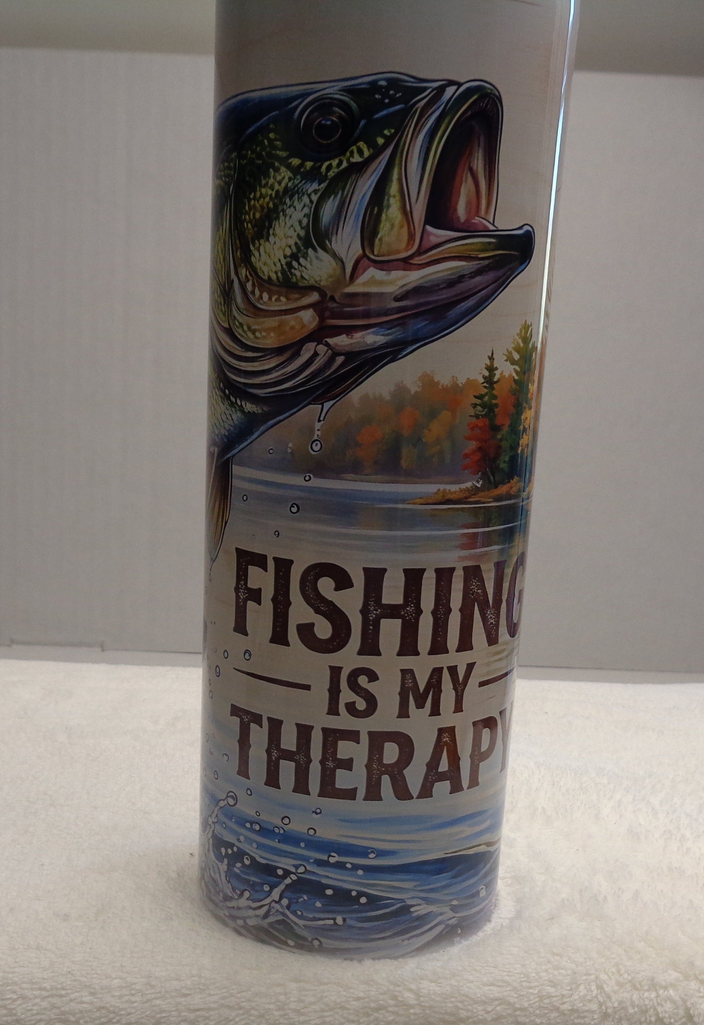 A cylindrical metal can with a fishing-themed label reading 'FISHING IS MY THERAPY' stands upright on a white towel. The label features a large bass fish illustration and blue water graphics.