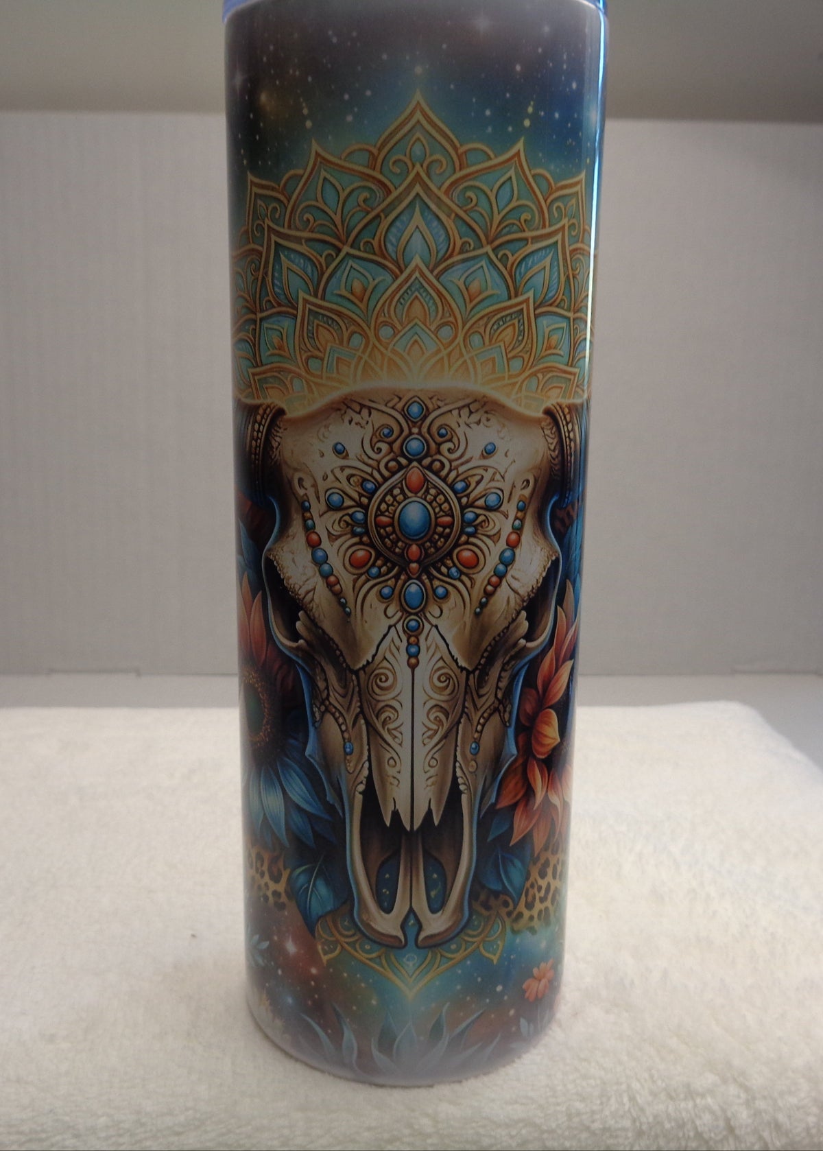 A tall cylindrical tumbler with a detailed graphic print of an elephant skull surrounded by mandala patterns and a starry sky, displayed on a white towel against a white background.