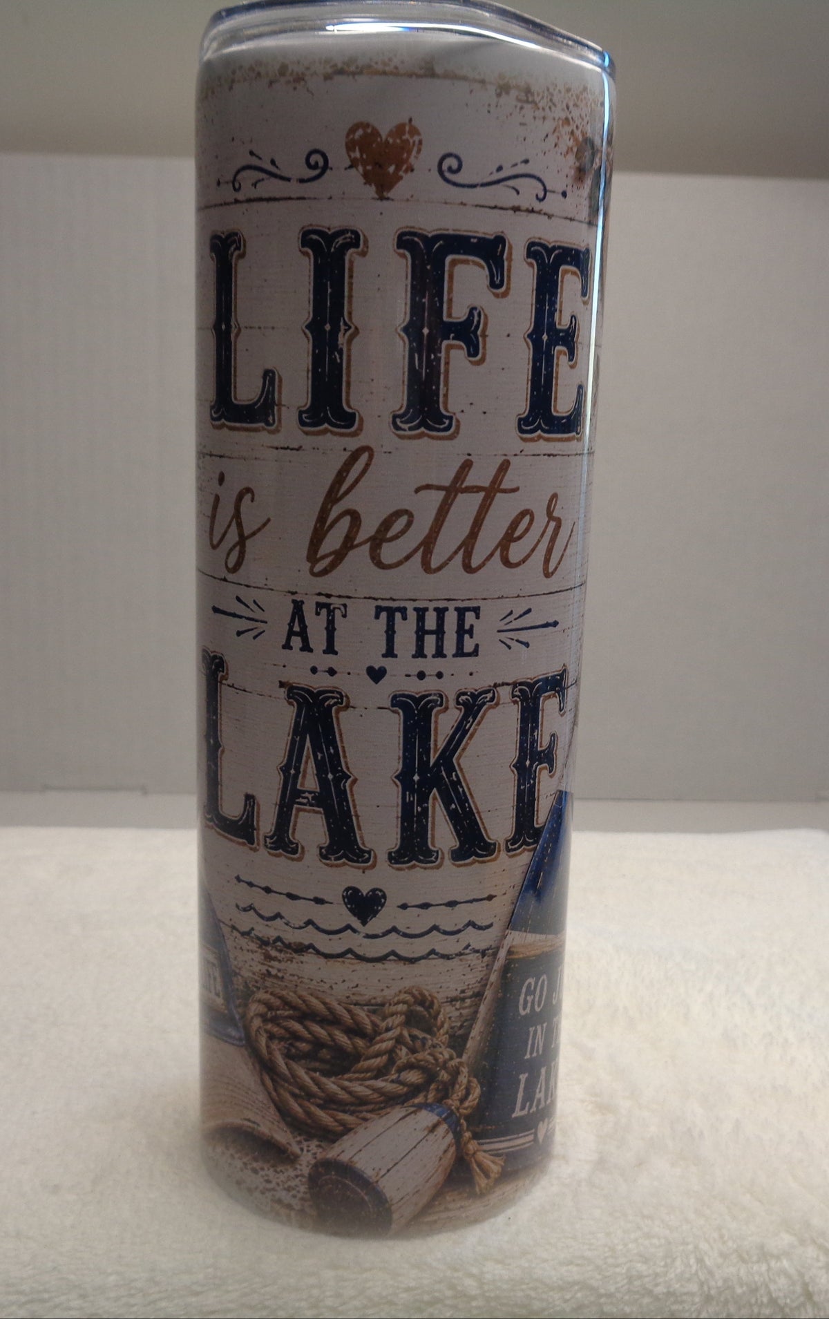 A cylindrical metal can with a rustic white label featuring the text 'LIFE is better AT THE LAKE' in navy and brown lettering, standing upright on a white towel.