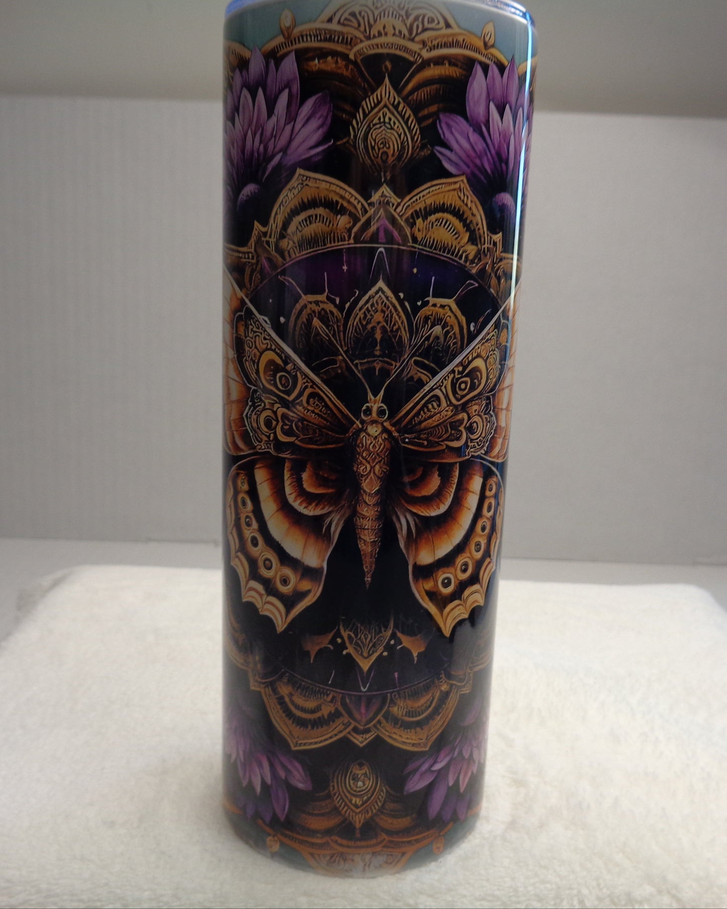A tall cylindrical tumbler with a detailed symmetrical butterfly and floral design in purple, gold, and black, sitting on a white towel against a white background.