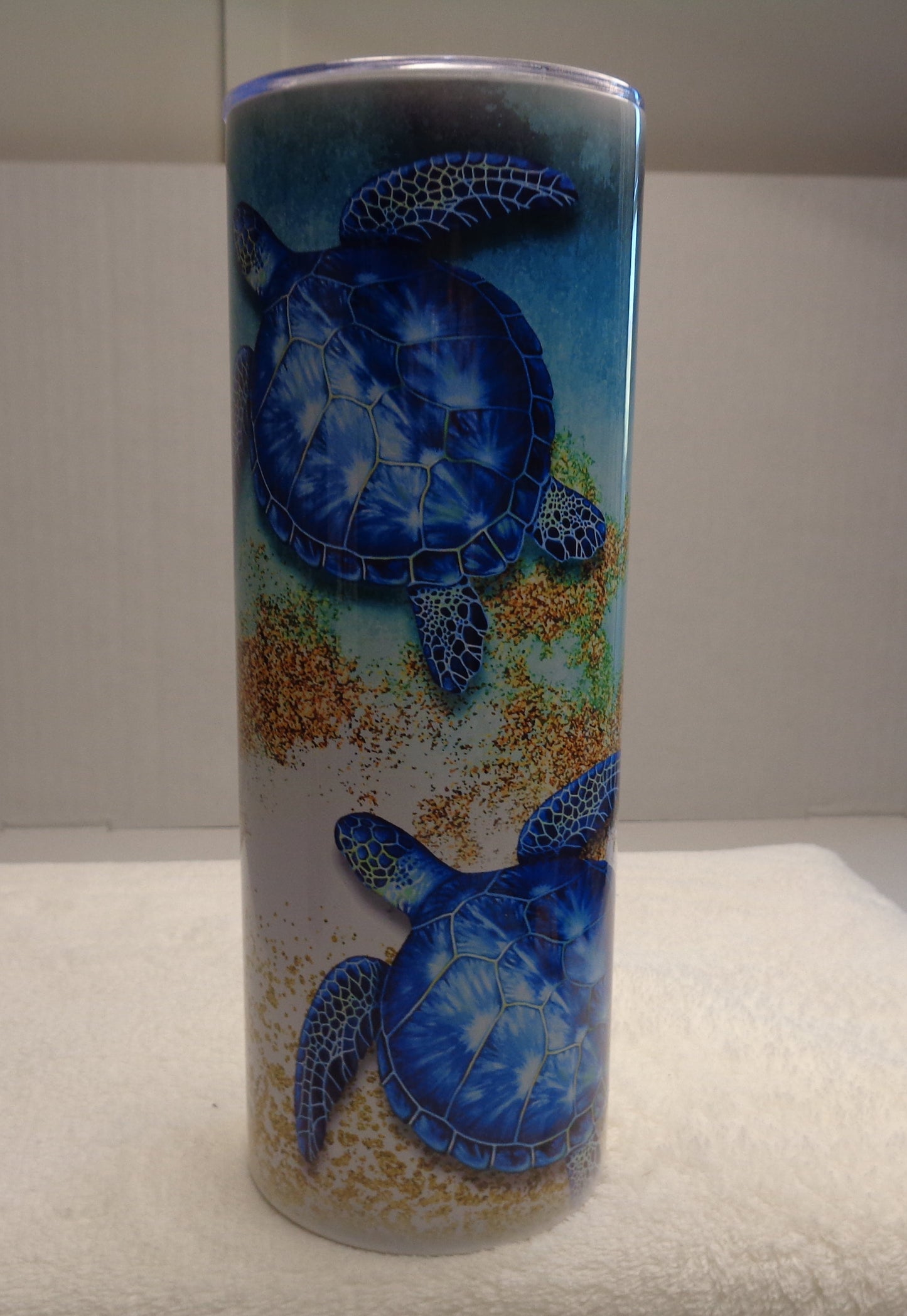 A tall cylindrical glass tumbler featuring a printed design of two blue sea turtles swimming over sandy ocean bottoms, displayed on a white towel in an indoor setting.