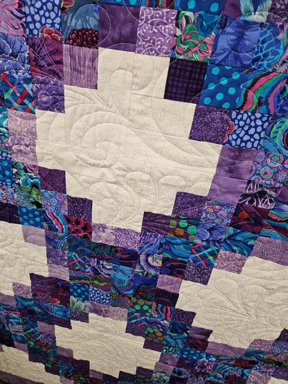 A close-up of a patchwork quilt featuring alternating blocks of purple-toned patterned fabrics and cream-colored fabric with swirling quilting.